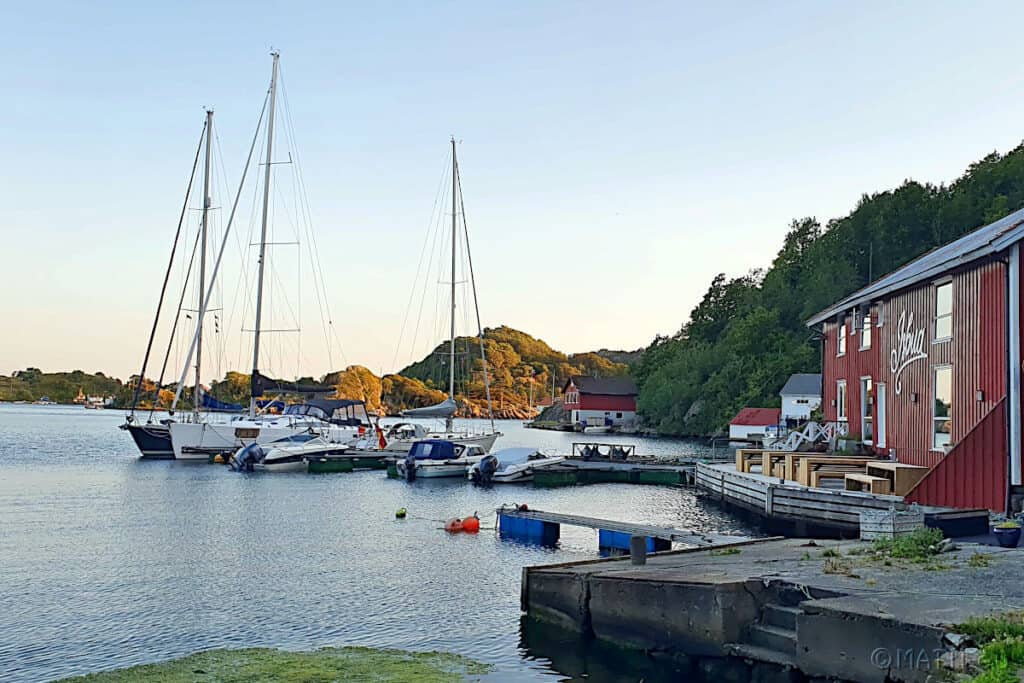 Yachthafen Kirkehamn mit Segelbooten und roten Bootsgebäuden am Wasser in schöner Küstenlandschaft.